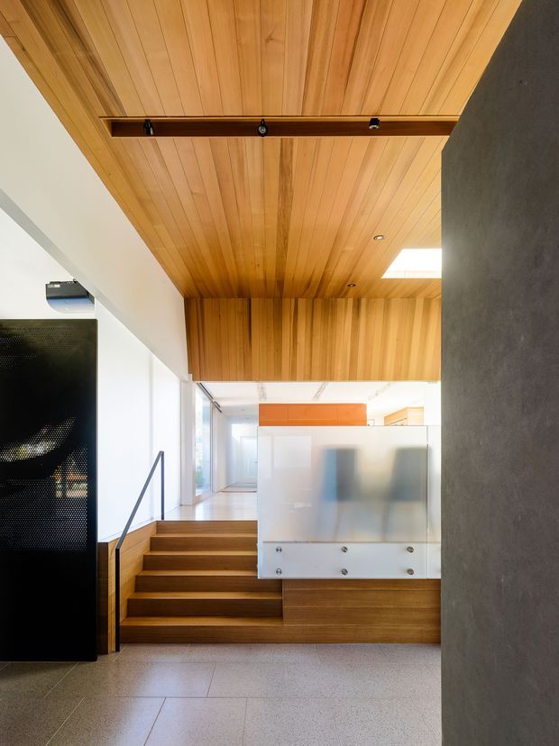 A contemporary interior circulation space with a timber-lined ceiling extending over a short flight of timber stairs. The stair landing includes a frosted glass privacy screen with visible fasteners, a dark textured wall, recessed and track-style lighting, and a large black perforated panel or screen. Light terrazzo-like tiled flooring and bright glazing create an open, refined architectural atmosphere.