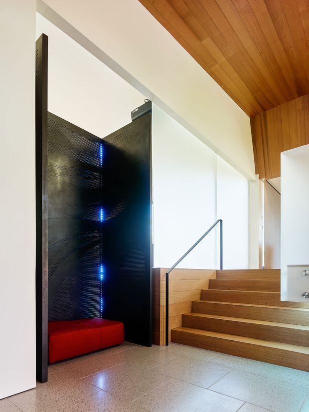 Interior view of a contemporary stairway with broad timber steps and a slim dark metal handrail. The stair rises beside a dramatic perforated black-metal feature screen with blue accent lighting and a built-in red upholstered bench. White walls, a warm timber-lined ceiling, and a polished light terrazzo or concrete-like floor create a crisp, high-contrast composition.