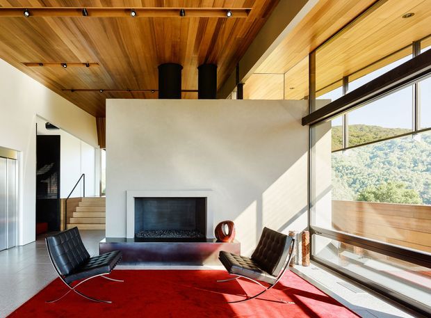 A bright modern living room with a full-height glazed wall overlooking a wooded hillside. The space features a dramatic white plaster fireplace wall, dark cylindrical chimney elements, extensive warm timber ceiling lining, a red area rug, and two black leather lounge chairs. A short stairway leads upward behind the fireplace wall.