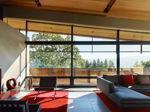 A bright, contemporary living room with expansive black-framed floor-to-ceiling glazing opening to a balcony or terrace with timber privacy screening and views of trees and distant hills. The room features a warm timber-lined ceiling with exposed structural beams, pale walls, large light-grey floor tiles, a dark sectional sofa with red cushions, and a low black lounge chair over a bold red rug. Strong sunlight creates crisp shadows across the space.