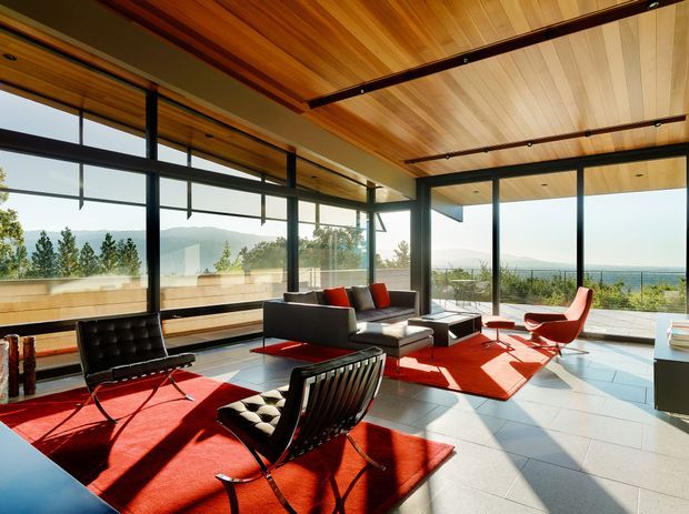 A bright, spacious modern living room with floor-to-ceiling black-framed glazing overlooking a coastal landscape. The room has a warm timber-lined ceiling with dark exposed beams, pale tiled flooring, sculptural mid-century-style lounge furniture, and vivid red area rugs and upholstery. Strong daylight creates a high-contrast, inviting interior.