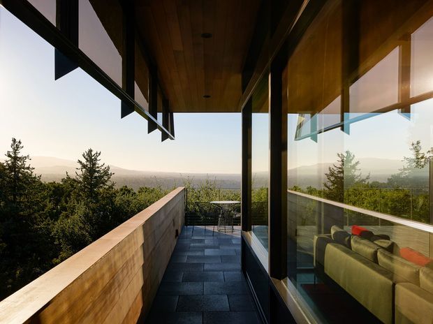 A narrow elevated balcony or terrace runs along a modern glass-walled building, overlooking a wooded landscape and distant hills. The space has dark large-format tiled paving, a warm timber soffit, black-framed floor-to-ceiling glazing, and a timber-clad parapet. Reflections and views through the glazing reveal a living room with a sofa inside. The image has a calm, warm modern architectural character.
