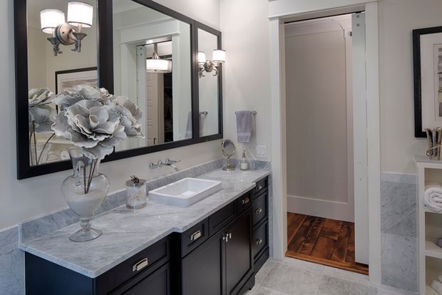 A refined bathroom vanity area with a long double-sink-style marble-look countertop, white vessel basin, dark built-in cabinetry, large black-framed mirror, wall sconces, and light grey wall and floor finishes. A doorway opens to a hall with dark timber flooring, while open shelving with neatly folded towels is visible at right.