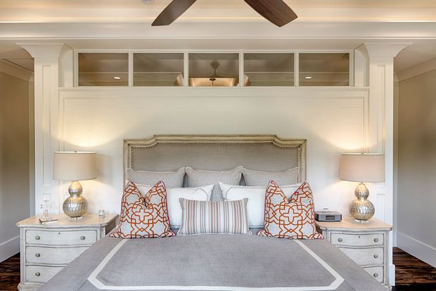 A refined traditional bedroom with a large upholstered bed, layered decorative pillows, matching bedside tables and lamps, white panelled walls, and clerestory-style interior glazing above the bed. The room has warm neutral tones with dark timber flooring and symmetrical built-in architectural detailing.