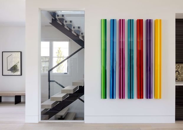 Bright contemporary interior featuring a sculptural open staircase with dark stringers and pale floating treads, viewed through a tall interior opening. A large wall-mounted arrangement of vertical multicoloured glossy rods provides a vivid feature installation, while pale walls and flooring create a clean, gallery-like setting. A partial bench and framed artwork are visible in the adjoining space.