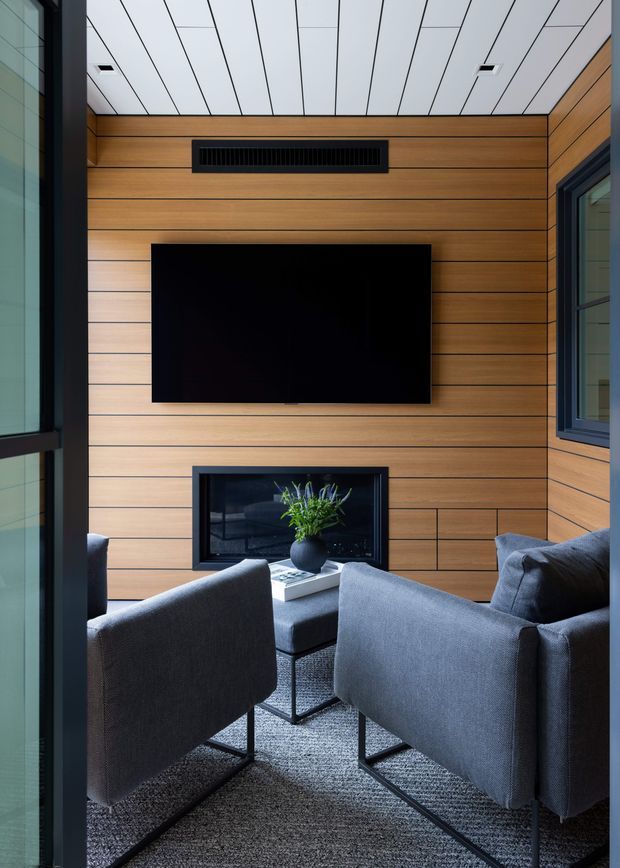 Compact contemporary media room with a warm horizontal timber-panelled feature wall, wall-mounted black television, linear air vent, and a low built-in fireplace. Four dark grey upholstered lounge chairs surround a small square coffee table, with large dark-framed windows and a white linear-panel ceiling. The room has a refined, cozy, modern feel.