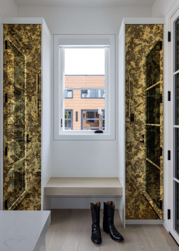 A bright, compact dressing and entry area with tall built-in wardrobes on both sides, finished in dramatic brown-gold veined panels with black hardware. A central window sits above a built-in bench, with pale timber flooring, white walls, and a partial marble-topped surface in the foreground. The space appears carefully fitted out and contemporary, with boots placed beneath the bench.