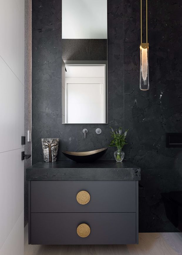 A compact, sophisticated bathroom vanity area with a dark textured stone-look wall and matching countertop. A matte black floating vanity has two drawers with round brass-gold pulls, supporting a sculptural black vessel basin with a brushed-gold inner surface. A frameless rectangular mirror, minimalist wall-mounted tapware, and a slender gold pendant light complete the dramatic, luxe composition. Pale timber flooring and a partially visible white door provide contrast.