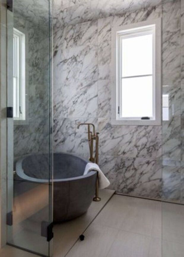 A refined bathroom featuring a freestanding dark grey bathtub beside a clear glass shower enclosure. The walls are clad in dramatic white-and-grey veined marble, complemented by a light neutral tiled floor, a white-trimmed window, and a freestanding brass-toned bath filler. A towel is draped over the tub, adding a small lived-in detail.
