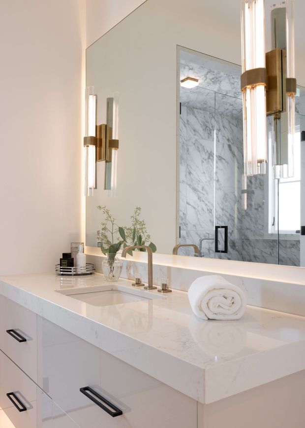 A bright, refined bathroom vanity with a large frameless mirror, white engineered-stone countertop, integrated rectangular basin, pale cabinetry, brushed brass gooseneck tapware, and contemporary vertical brass-and-glass wall sconces. The mirror reflects a marble-look tiled shower enclosure. The palette is predominantly white and light grey with warm metallic accents.