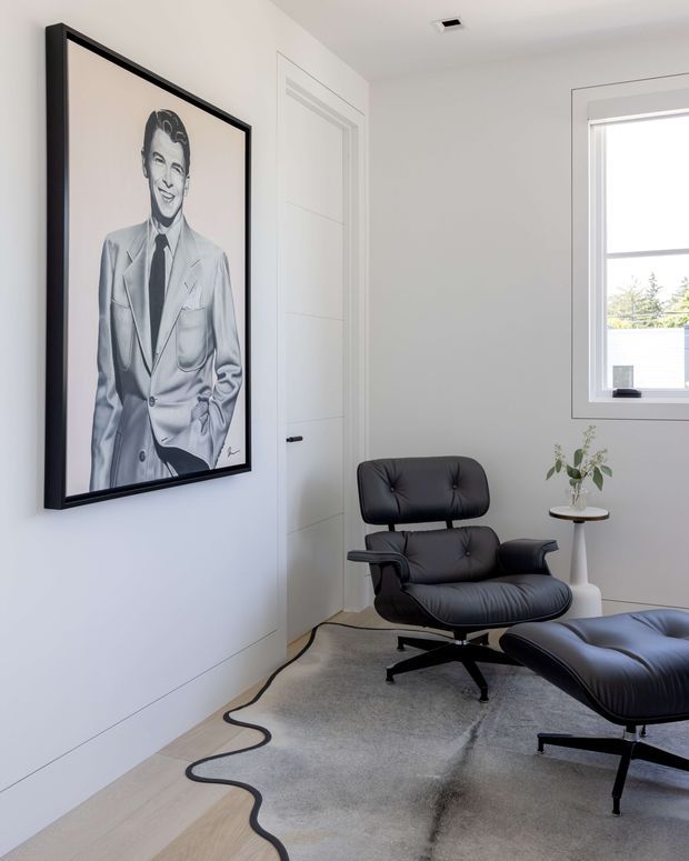 A bright, minimalist sitting room with white walls, a concealed white door, pale timber flooring, and a large neutral rug with a black organic-shaped border. A black leather lounge chair and matching ottoman sit beside a small round side table, while a large monochrome portrait in a black frame forms the main wall feature. A window provides natural light and views of greenery outside.