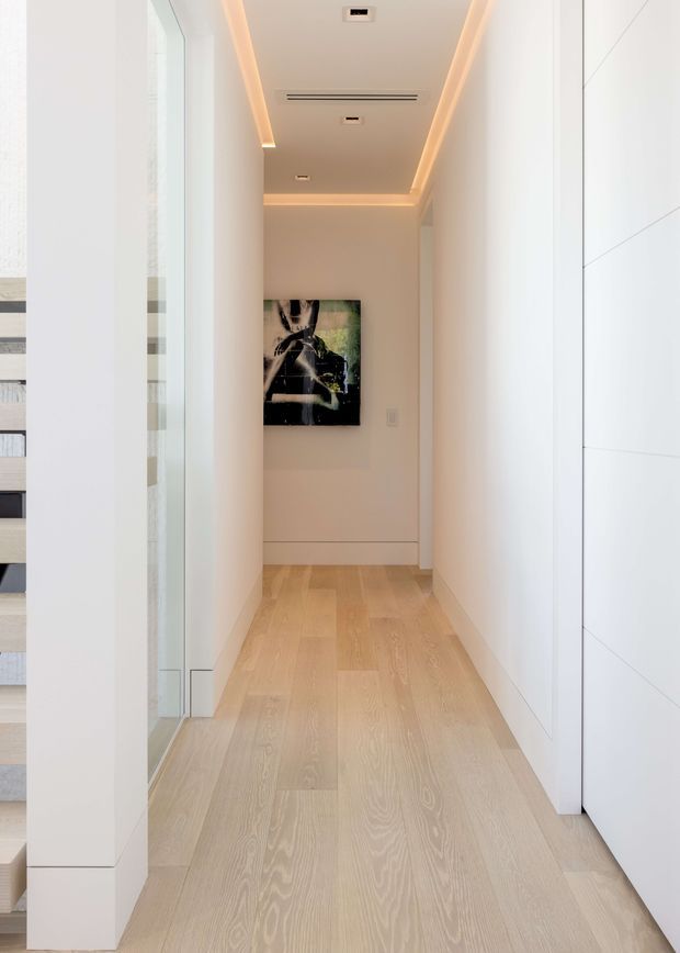 A narrow, bright residential hallway with pale timber plank flooring, white walls, and full-height white built-in cabinetry on the right. A glazed opening and shelving are visible on the left, while recessed linear ceiling lighting with warm concealed illumination leads toward abstract artwork at the far end.