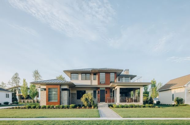 Front exterior view of a large two-storey contemporary suburban residence with a low-pitched metal roof, brick and light rendered wall sections, extensive glazing, timber-toned vertical cladding accents, covered entry and upper-level terrace. The home is set behind a manicured lawn with a straight paved path and structured garden beds.
