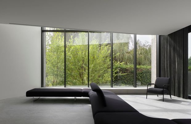Minimalist contemporary living room with a low black sectional sofa and matching chaise arranged around expansive floor-to-ceiling glazing. A sculptural black lounge chair sits near the window, while dark vertical drapery or slatted wall treatment frames the right side. The room has a smooth light-grey floor, white ceiling and walls, abundant natural light, and a leafy garden view beyond the glass.