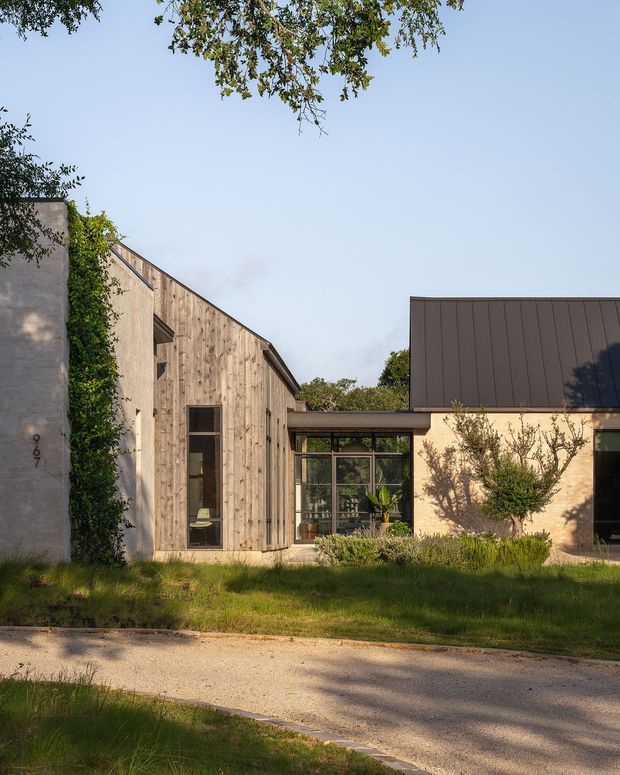 Street-facing view of a low-rise contemporary residence composed of several connected volumes. The facade combines vertically oriented weathered timber cladding, pale rendered or concrete walls, dark standing-seam metal roofing, and extensive glazed connector windows. Mature landscaping and a softly planted front verge frame the house in warm afternoon light.