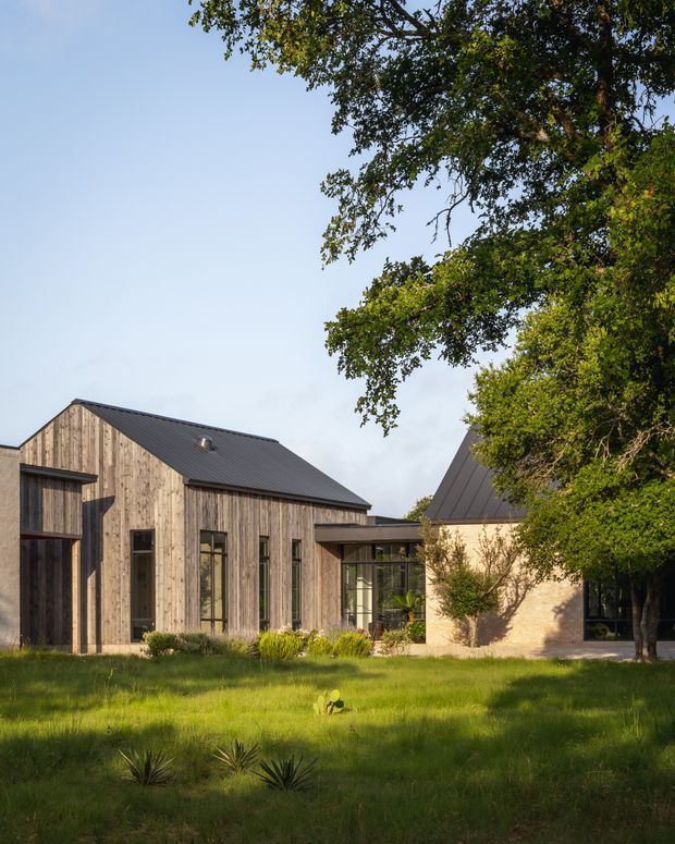 Exterior view of a contemporary multi-volume residence set in a landscaped garden. The building combines vertical natural timber cladding, dark standing-seam pitched roofs, expansive dark-framed glazing, and a pale masonry volume. Mature trees and lawn surround the house, with warm late-day sunlight creating a calm residential setting.