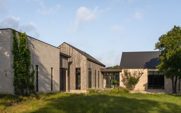 Exterior view of a low-rise contemporary building complex arranged around a landscaped courtyard. The composition combines pale textured masonry walls with vertical timber cladding, dark metal-framed glazing, and steep pitched metal roofs. Mature trees, climbing greenery, and lawn soften the restrained architectural forms.