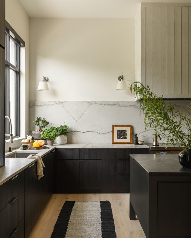 A refined kitchen with dark timber cabinetry, a large central island, pale stone-look splashback, light timber flooring, and tall steel-framed windows. Wall-mounted sconces, potted greenery, and restrained artwork add warmth and texture. The space has a narrow galley-like layout opening around the island, with a calm, contemporary feel.