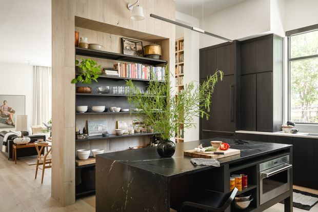 A bright, refined contemporary kitchen with a large dark stone island, black built-in cabinetry, open timber shelving, and integrated appliances. The island includes seating and a styled surface with a vase and serving items. A living area is partially visible beyond the kitchen, while large windows bring in abundant natural light.
