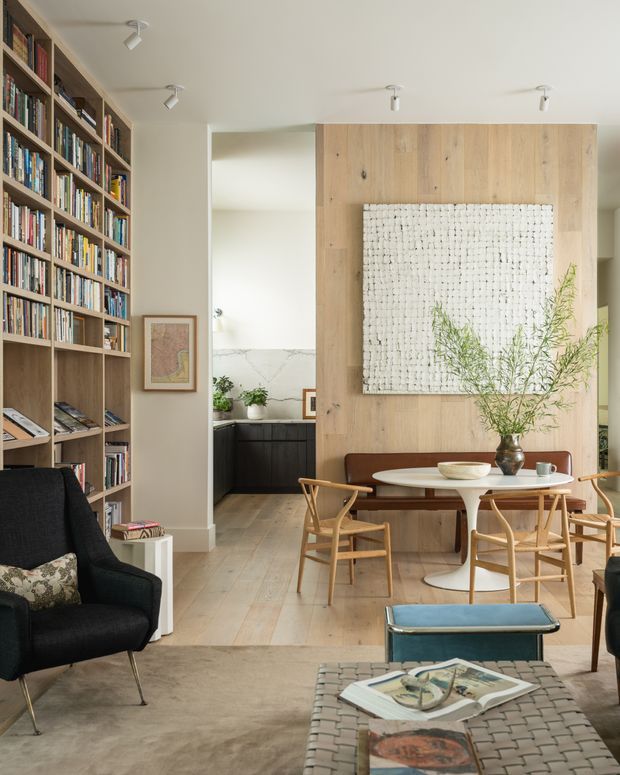 Warm, refined open-plan living and dining area with a full-height built-in bookcase, pale timber feature wall, textured artwork, and a compact kitchen visible beyond. A round white dining table with timber chairs sits in front of a brown upholstered banquette, while a dark lounge chair and woven coffee table occupy the foreground. Natural materials, soft neutral tones, and abundant books create an inviting contemporary interior.