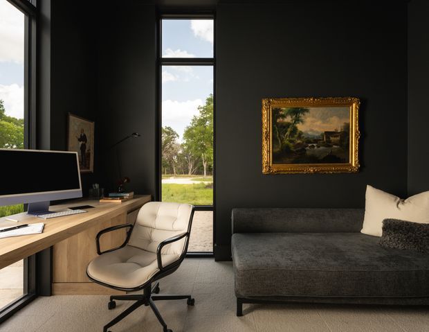 A sophisticated dark-toned home office and lounge corner with floor-to-ceiling black-framed glazing, a timber desk and computer setup, a cream swivel office chair, and a charcoal sofa with cushions. A traditional landscape painting in an ornate gold frame provides a contrasting decorative feature against the matte black wall, while the glazing opens views to greenery outside.