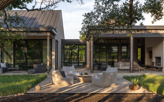 A contemporary single-storey residence arranged around a landscaped outdoor living area. Large glazed walls connect the interior living spaces to a paved courtyard-style terrace furnished with sculptural lounge chairs and a low fire pit or table. The building has restrained rectangular volumes, dark metal-framed glazing, a standing-seam metal roof, exposed timber or concrete-toned posts, and a visible interior fireplace. Mature trees and lawn surround the terrace.