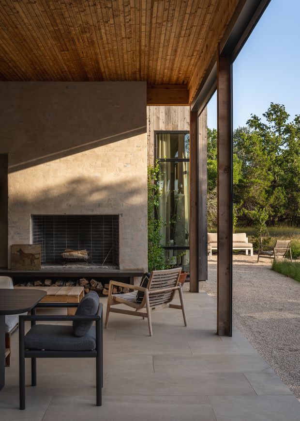 A refined indoor-outdoor living space with a large built-in open fireplace set into a textured pale stone or plaster wall. The covered terrace has a warm timber slatted ceiling, dark slender structural posts, expansive glazing, and a pale large-format tiled floor. Lounge chairs and a round table sit beside the fireplace, opening onto a gravel courtyard with outdoor seating and mature vegetation.