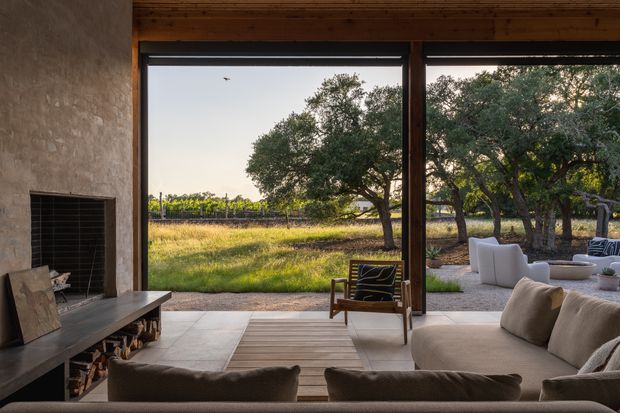 A warm, rustic-modern living room opening directly onto a landscaped outdoor seating area and vineyard-like rural landscape. The room features a large masonry fireplace with built-in firewood storage, full-height dark timber-framed glazing, a timber-lined ceiling, pale tiled flooring, and a large neutral sectional sofa. Outdoor lounge furniture and mature trees are visible beyond the glazing.