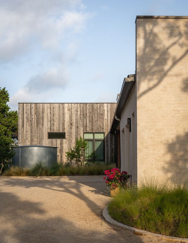 Exterior view of a contemporary residential or small-scale architectural building, showing a tall rendered masonry wall on the right, a weathered vertical timber-clad volume with dark-framed windows at the rear, and a corrugated metal cylindrical water tank. A gravel driveway and curved concrete edging lead through low ornamental grasses and planting. Warm late-afternoon light casts tree shadows across the facade and ground.