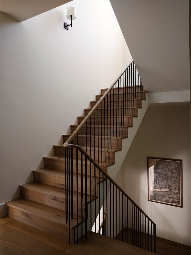 Interior view of a contemporary stairway with warm timber treads and risers, slim black metal vertical balustrades, and light painted walls. The stair turns around a central landing, with a wall-mounted light fixture and framed artwork visible on the stairwell walls. The space is softly lit and double-height or visually open above the stairs.
