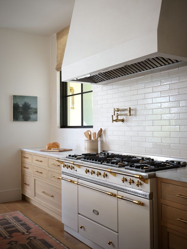 A refined kitchen centred on a large white professional-style range cooker with brass controls and hardware, beneath a substantial white rangehood. The backsplash is composed of glossy white subway tiles with light grout, flanked by pale painted cabinetry on the left and darker timber cabinetry on the right. A black-framed window, timber flooring, brass pot filler, utensil crock and patterned runner complete the warm, classic interior.
