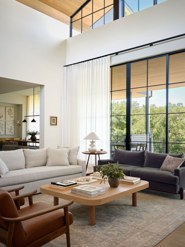 A bright, refined double-height living room with a large seating arrangement around a low timber coffee table. The room features floor-to-ceiling black-framed glazing and glass doors opening to a balcony or terrace, white walls, a warm timber-lined ceiling and upper clerestory glazing, sheer curtains, neutral sofas, leather lounge chairs, and layered soft furnishings. A dining area is partially visible through the opening at left.