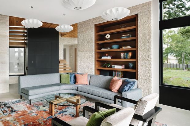 Contemporary living room with a large grey L-shaped sectional sofa, sculptural white pendant lights, a built-in dark timber bookcase set into a textured stone feature wall, and a black-framed glazed door or window. The room includes a patterned area rug, glass coffee table, and timber ceiling detailing, with a bright outlook to greenery outside.