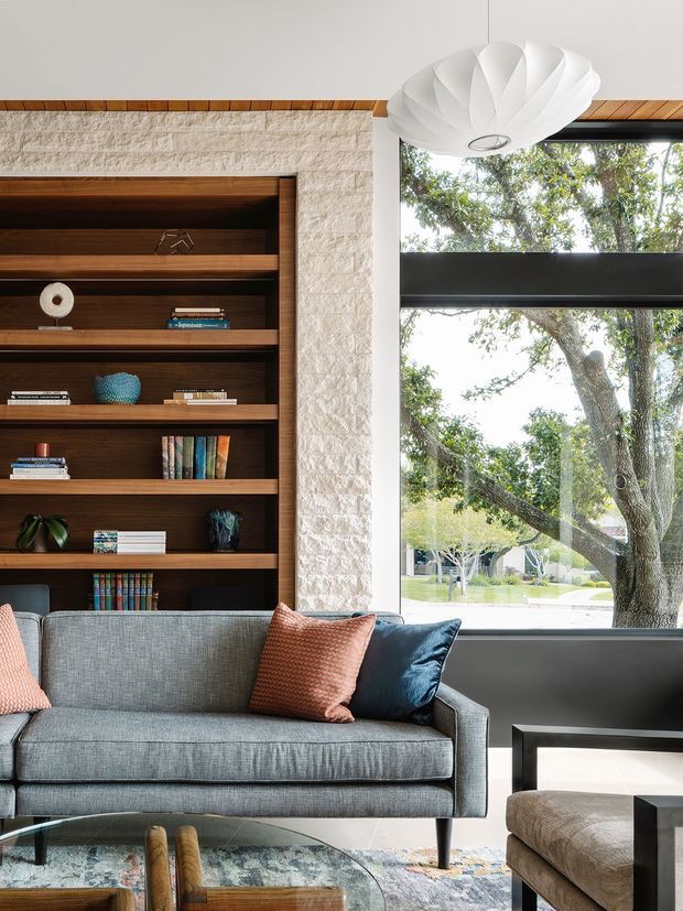 A bright, refined living room with a grey upholstered sofa positioned in front of dark timber built-in shelving. The shelving is set into a textured light stone wall and displays books and decorative objects. A large window provides views of mature trees and brings in abundant daylight. A sculptural white pendant light hangs above, with a dark side table, patterned rug, glass coffee table, and blue and coral cushions completing the room.