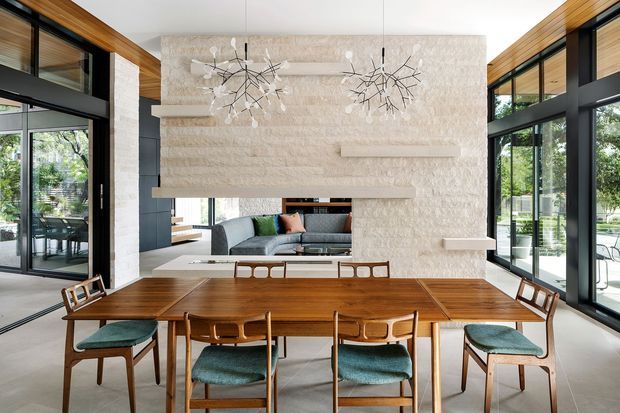 Bright, double-height contemporary dining and living space with a long mid-century-style timber dining table and upholstered dining chairs in the foreground. A large textured pale stone feature wall with integrated shelving and a fireplace-like opening separates the dining area from the lounge. Extensive black-framed floor-to-ceiling glazing, warm timber ceiling and wall detailing, sculptural pendant lights, and views to greenery create an airy indoor-outdoor connection.