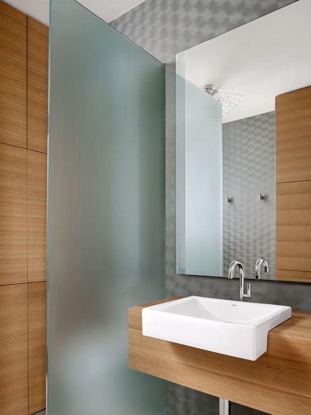 Contemporary bathroom vanity with a white rectangular vessel basin, chrome wall-mounted mixer, warm timber cabinetry and benchtop, patterned grey wall and ceiling surfaces, and a large frameless mirror. A frosted glass privacy screen occupies the left side of the room.