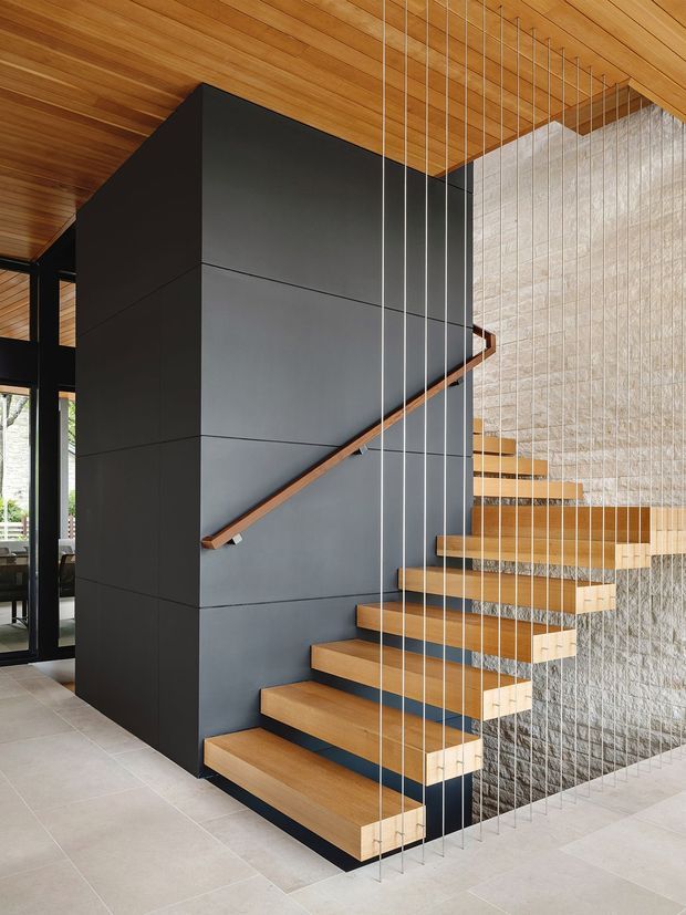 Contemporary interior stairway with floating timber treads cantilevered from a dark charcoal-clad stair wall. Vertical metal safety cables form a balustrade, while a warm timber-lined ceiling and pale stone-textured wall create contrast. Glazing and a living area are visible beyond the stair enclosure.