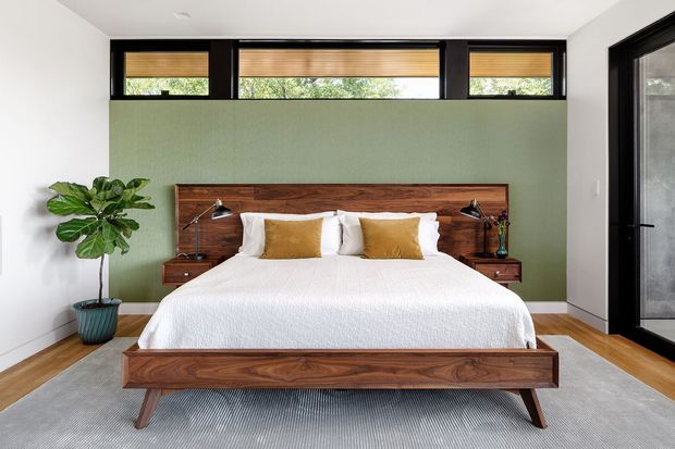 A bright, modern bedroom centred on a low-profile walnut platform bed with an integrated wood headboard and matching bedside shelves. White bedding and walls contrast with a muted sage-green feature wall, black-framed clerestory windows, and a black-framed glazed door. A large grey rug, warm timber flooring, bedside lamps, and a potted fiddle-leaf fig complete the calm, contemporary composition.