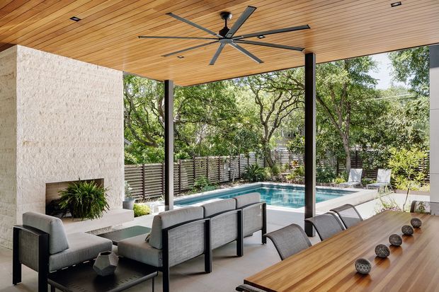 A sophisticated covered outdoor entertaining area beside a swimming pool, featuring a timber-lined ceiling with a large ceiling fan, dark structural posts, a textured stone fireplace wall, contemporary outdoor lounge seating, and a long timber dining table. The landscaped private courtyard and pool are visible beyond the open sides.