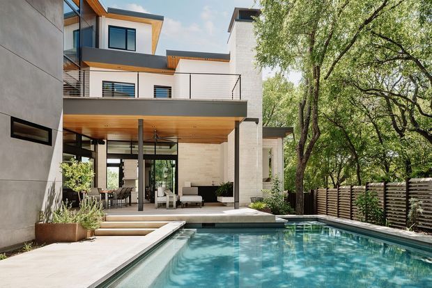 Exterior view of a contemporary multi-level residence with a private swimming pool in the foreground. The house features clean white rendered and stone-clad walls, dark-framed glazing, projecting flat roof forms, slim metal balustrades, and a warm timber-lined covered terrace ceiling. The pool terrace opens to a landscaped garden enclosed by a horizontal slatted privacy fence.
