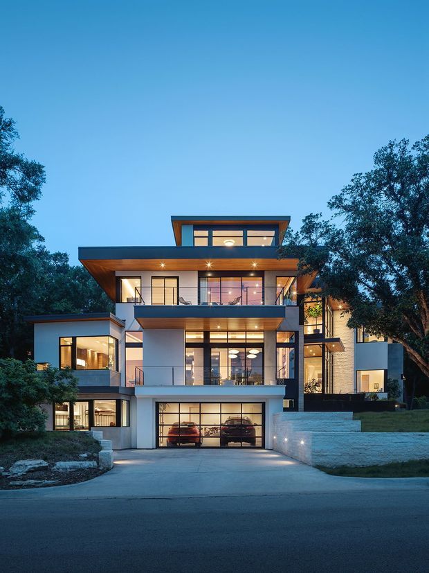 Twilight exterior view of a large contemporary multi-storey residence set among mature trees. The house features strong horizontal roof overhangs, extensive floor-to-ceiling glazing, warm timber soffits, pale rendered and masonry walls, multiple terraces and balconies, and an integrated double garage accessed by a broad driveway. Interior lighting creates a warm glow through the windows.