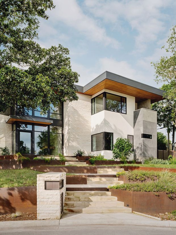 Street-facing photograph of a contemporary two-storey residence set on a landscaped sloping site. The house features strong geometric volumes, extensive dark-framed glazing, pale textured masonry walls, a projecting flat roof with a warm timber-lined soffit, and a sculptural concrete entry tower. Terraced retaining walls, garden planting, and a stepped pedestrian approach lead from the street to the entrance.