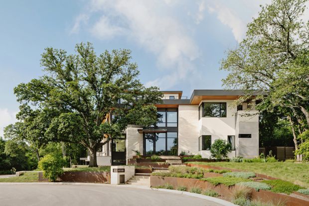Exterior view of a contemporary multi-level residence set among mature trees, with a pale rendered facade, extensive dark-framed glazing, projecting flat roof forms, landscaped terraced garden beds, and a street-facing entry. The composition highlights the building facade and its connection to the sloping landscaped site.