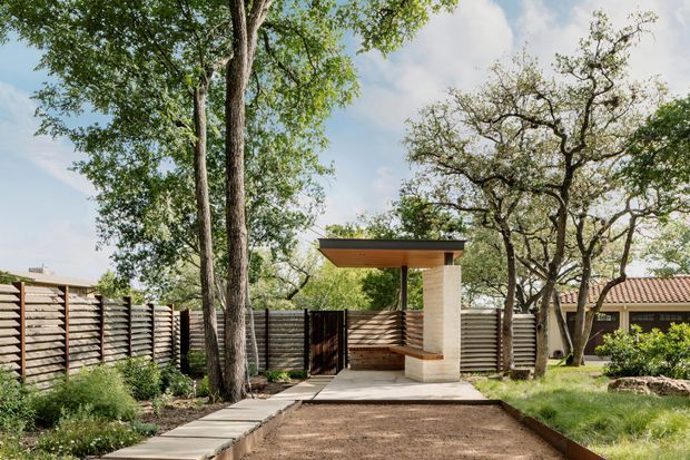 A landscaped residential courtyard featuring a modern covered outdoor structure or pavilion. The structure has a thin flat roof with a dark fascia, a light masonry or rendered column, and a built-in timber bench beneath it. A long horizontal timber privacy fence encloses the space, with mature trees, planted garden beds, a paved path, and a rectangular decomposed-granite or gravel area in the foreground. A tiled-roof house is partially visible behind the fence.