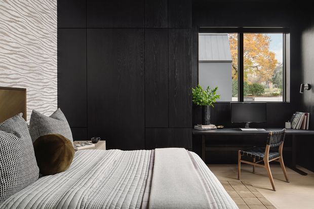 A moody, refined bedroom with a large upholstered bed in the foreground, dark full-height timber wall panelling and integrated joinery, and a compact built-in desk beneath a black-framed window. The room combines black and charcoal surfaces with a light textured feature wall, pale bedding, patterned cushions, and warm autumn views outside.