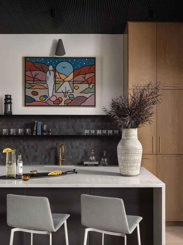A sophisticated built-in home bar and entertaining area with a pale stone counter, dark textured tile backsplash, open glassware shelving, brass faucet, timber cabinetry, and two upholstered bar chairs. A colourful framed artwork and sculptural vase add visual interest. The room has a dark slatted ceiling and warm, refined finishes.
