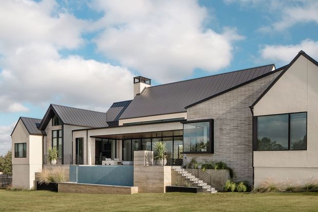 Exterior view of a large contemporary residence with multiple pitched gabled roof forms, pale rendered walls, dark metal roofing, and a textured grey masonry feature wall. Extensive glazing connects the interior to a raised terrace and swimming pool, with stairs descending to landscaped lawn.
