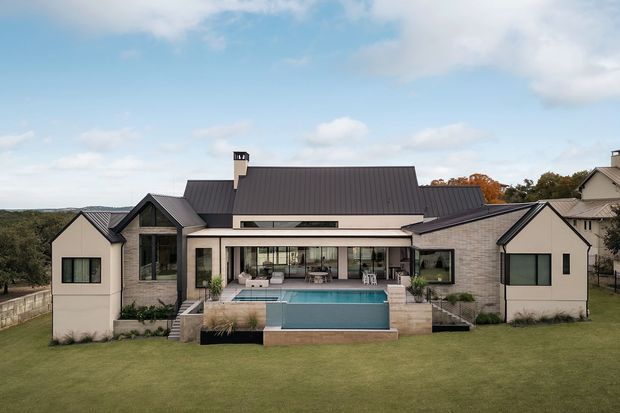 A large contemporary single-storey residence viewed from the rear, with a dark pitched metal roof, pale rendered and brick exterior walls, extensive glazed openings, and a covered outdoor entertaining terrace. The terrace overlooks a rectangular swimming pool and broad manicured lawn, with landscaped planting and surrounding trees visible beyond.