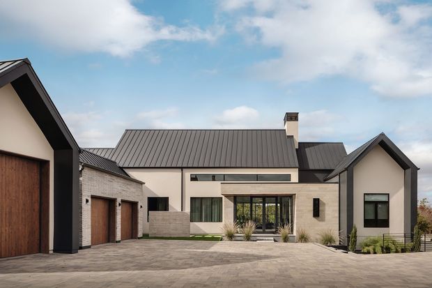 Contemporary single-storey residential home viewed from the front, arranged around a broad paved driveway or courtyard. The facade combines pale rendered walls, light masonry, dark standing-seam metal gabled roofs, dark window and door frames, and warm timber garage doors. Multiple connected gabled volumes frame a central glazed entry or living area, with restrained planting along the facade.