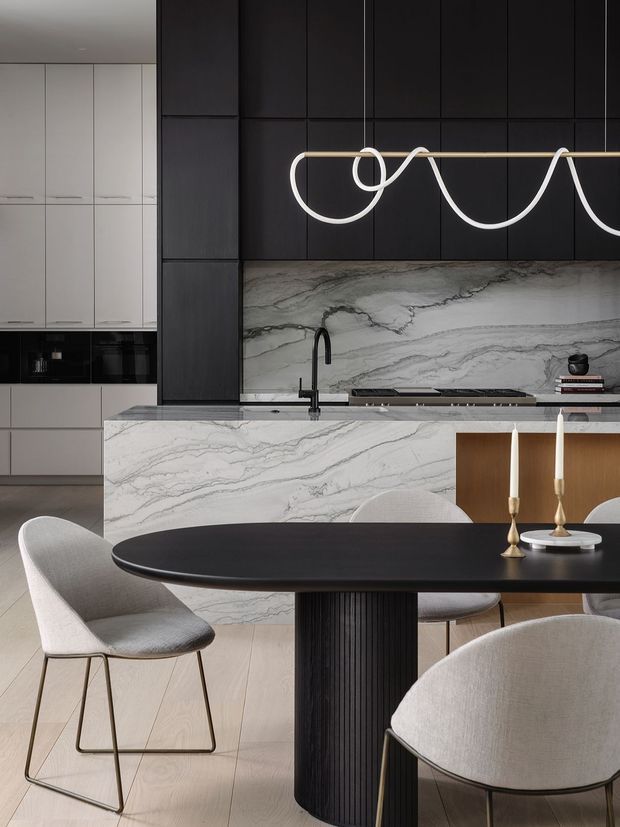 A refined open-plan kitchen and dining area with a dramatic veined stone island and backsplash, matte black full-height cabinetry, integrated appliances, and a sculptural linear pendant light. A dark oval dining table with a fluted black pedestal is surrounded by light upholstered dining chairs, while brass candlesticks provide a subtle accent. The space has a minimalist, high-contrast and luxurious contemporary feel.